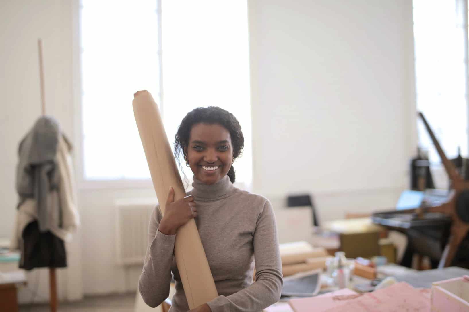 smiling young ethnic craftswoman with paper roll in workshop